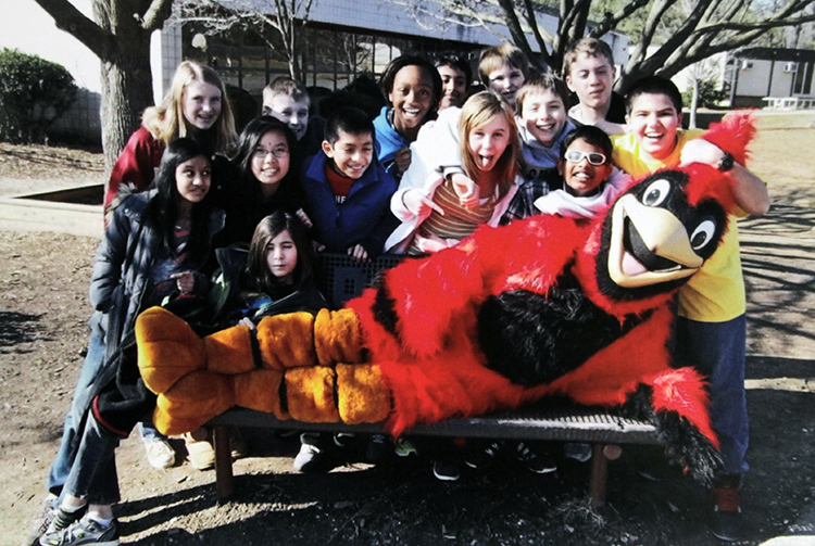 Photograph of Cherry Run’s cardinal mascot reclining on a bench in front of the school. The bench is surrounded by smiling students. 