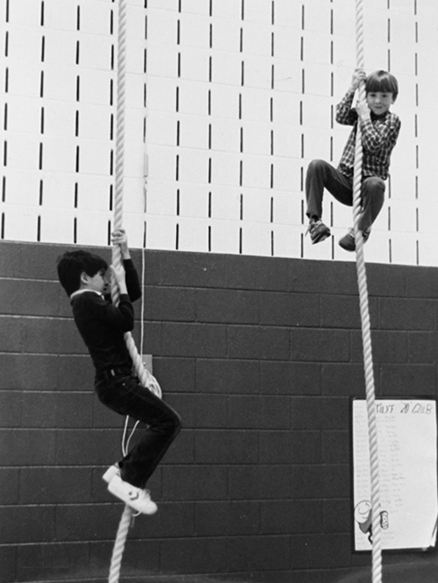 Black and white photograph of two students climbing ropes in the gymnasium.