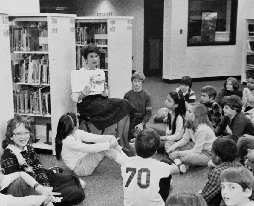 Black and white yearbook photograph of students sitting on the floor in the library. They are looking up at a teacher who is holding open a book and reading it to them.