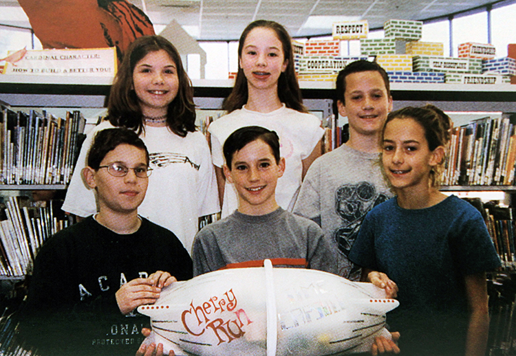 Yearbook photograph of six upper-grade students standing behind a time capsule. The capsule is approximately three feet long, is white in color, and is shaped like a football.